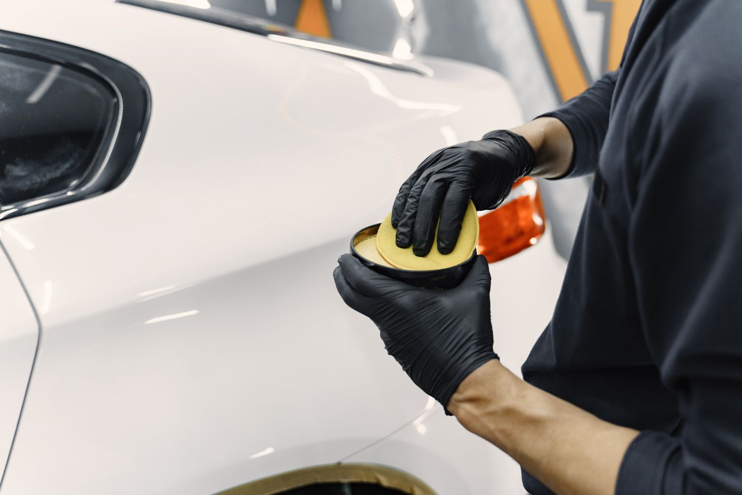A person in black gloves carefully applies car wax from a container using a yellow applicator to a white car