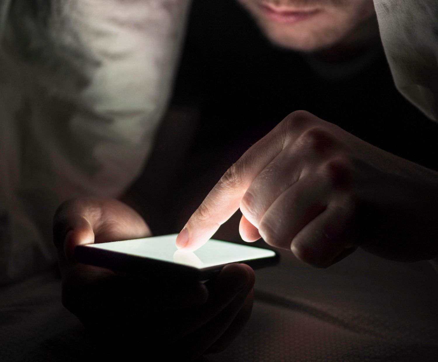 A person secretly uses a brightly lit smartphone under covers in a dark room