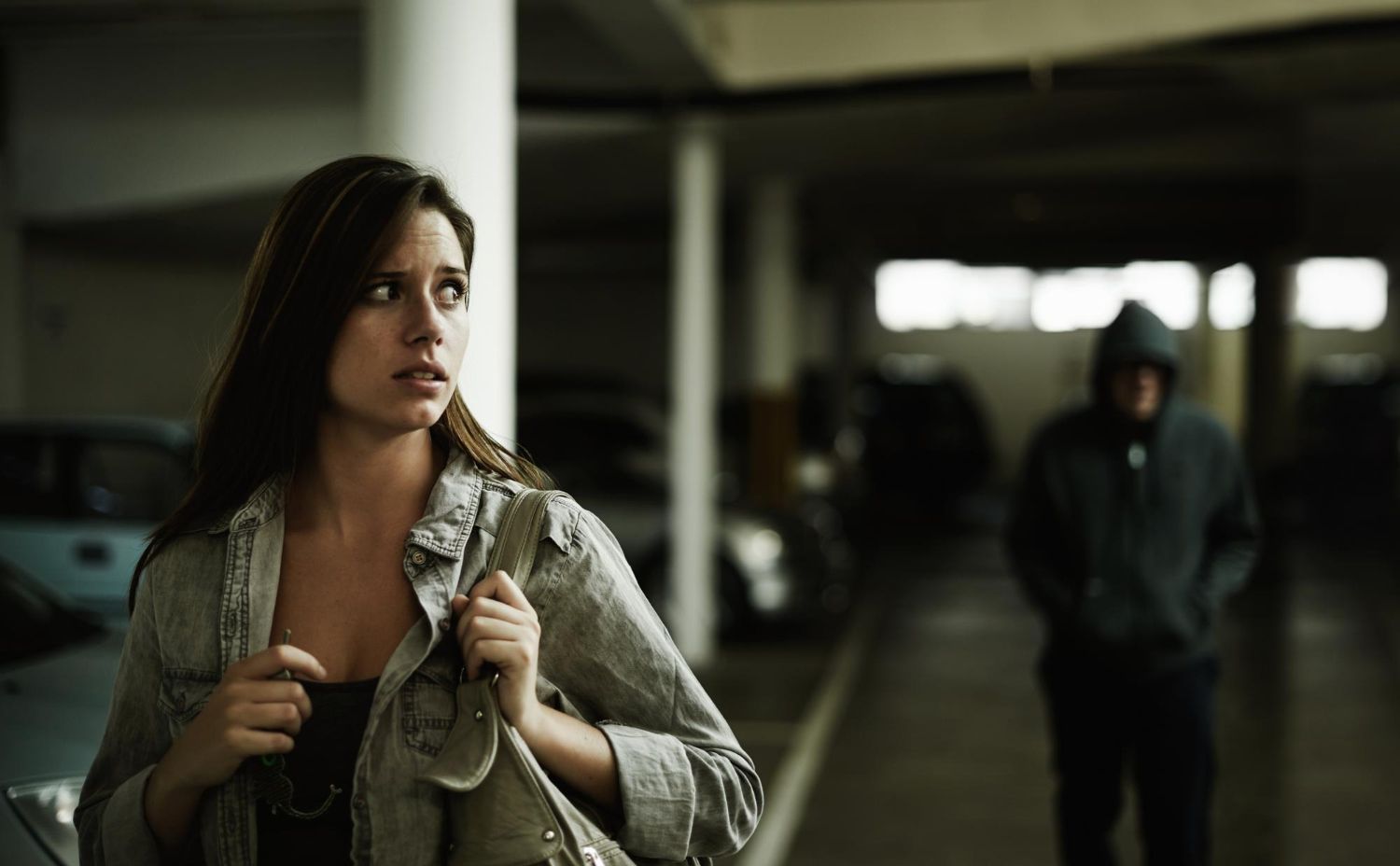 A fearful woman in a dimly lit parking garage looks over her shoulder at a lurking hooded figure in the distance