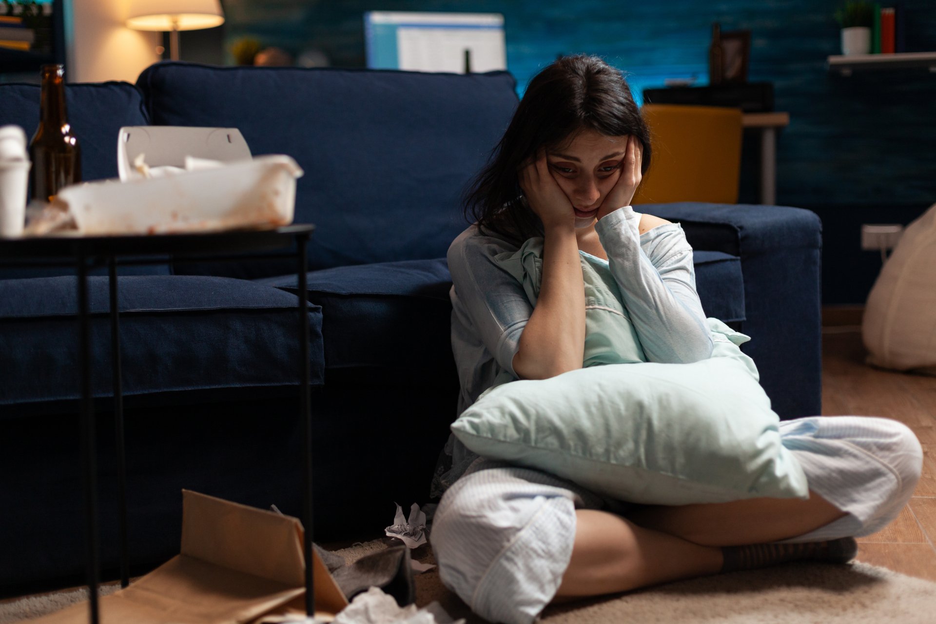 A distraught woman in pajamas sits on the floor amidst clutter, holding a pillow