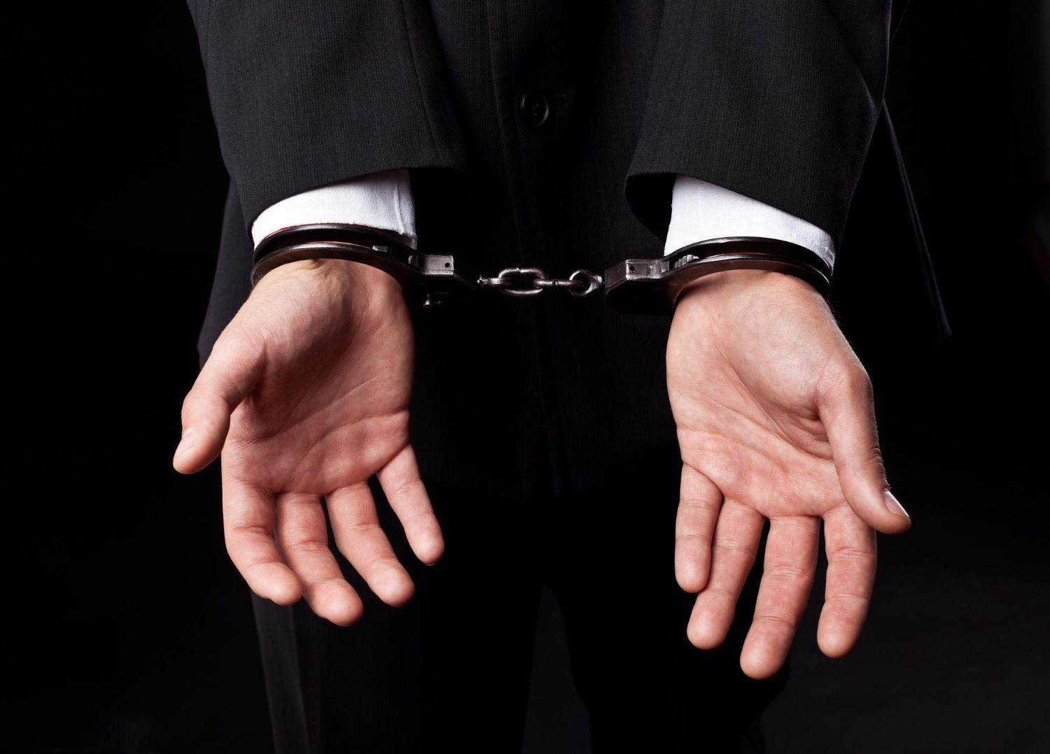 A person in a dark suit is shown from behind, hands cuffed at the wrists