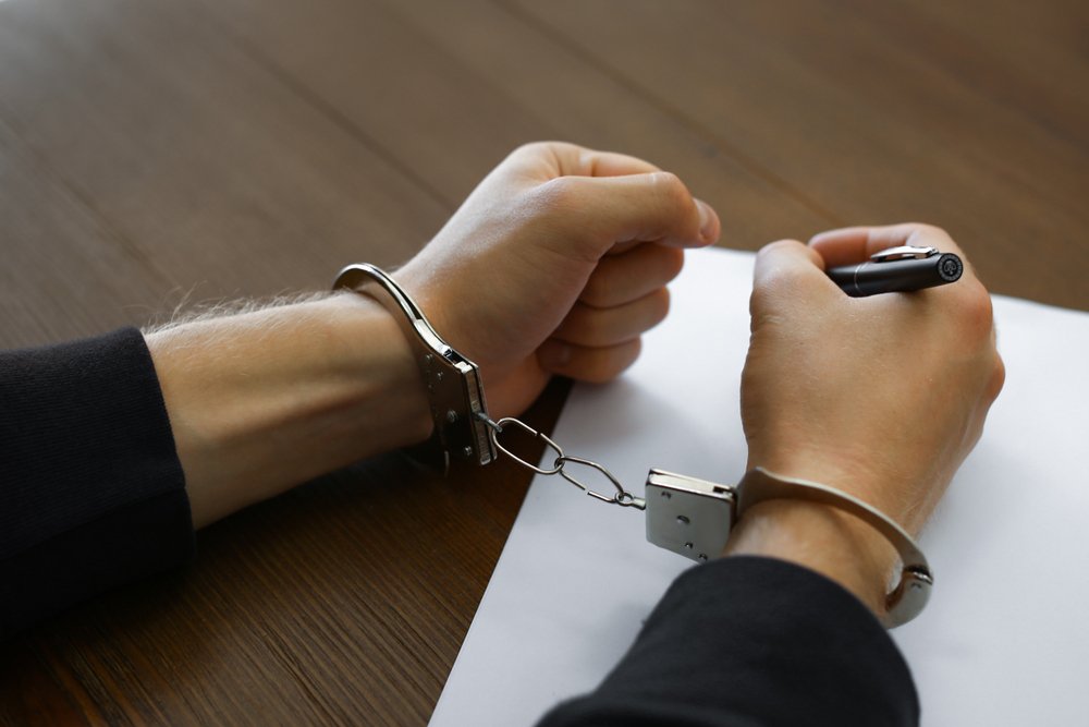 A close-up shows a person's hands handcuffed together at a table