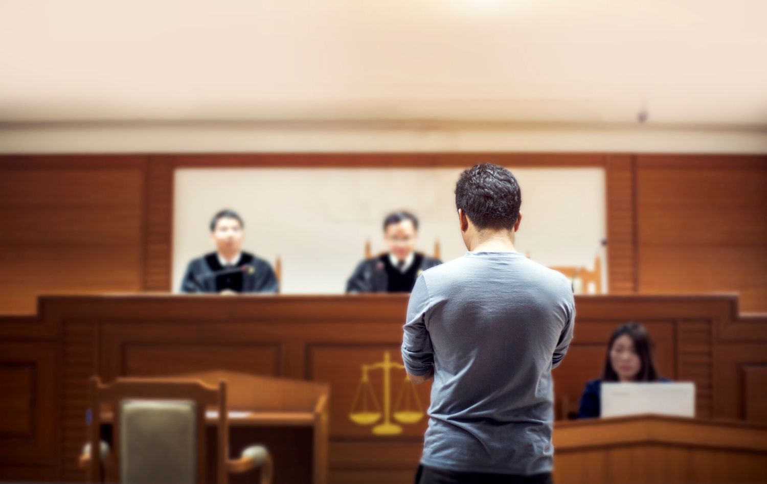 A man stands with his back to the viewer in a blurred courtroom, facing two judges