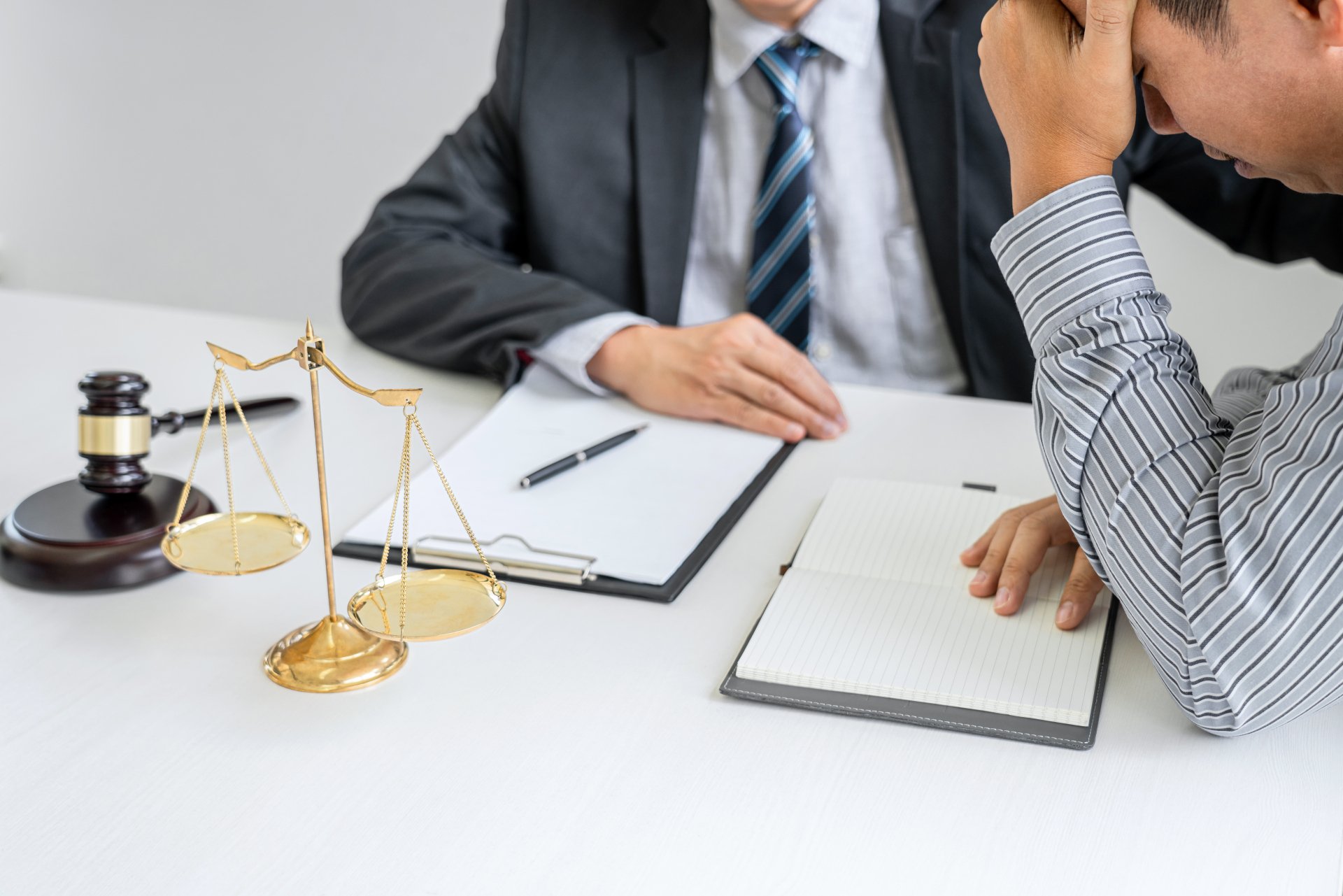 A lawyer in a suit consults with a distressed client across a desk with a scale of justice and gavel