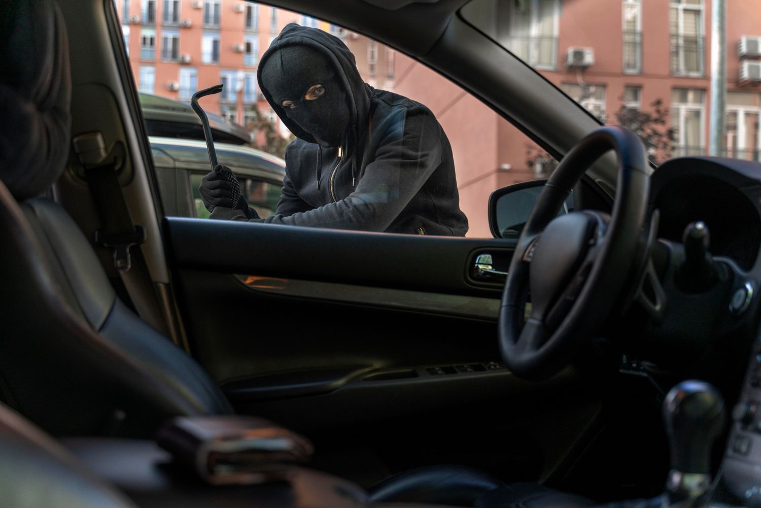 A masked figure with a crowbar attempts to break into a car