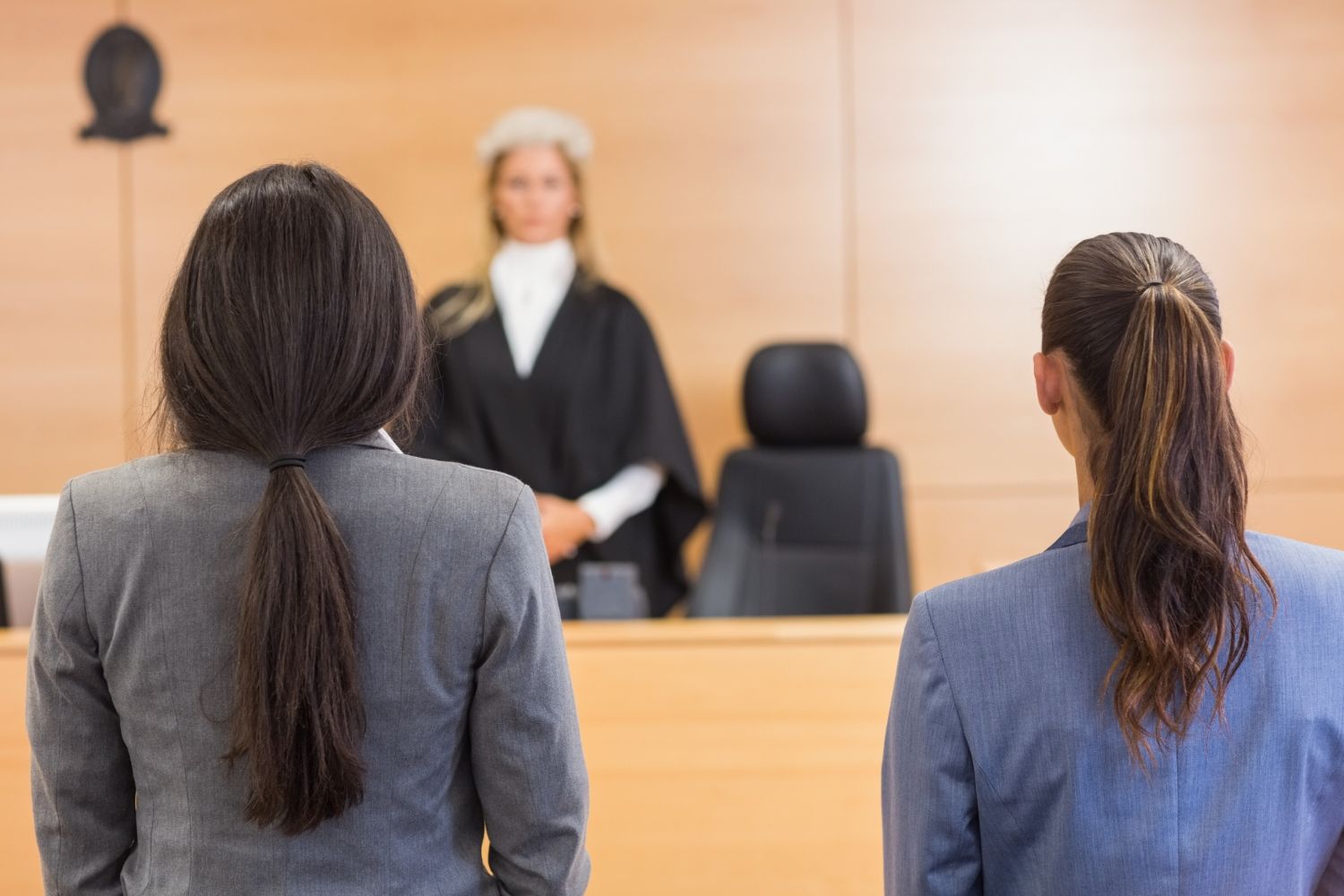 Two individuals in suits stand with their backs to the viewer, facing a robed judge in a formal courtroom