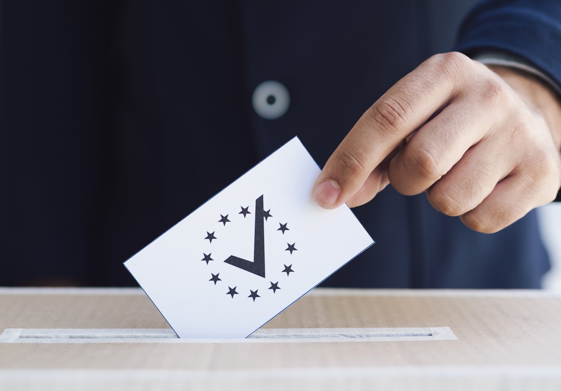 A hand places a ballot, featuring a large checkmark surrounded by twelve stars, into a voting box