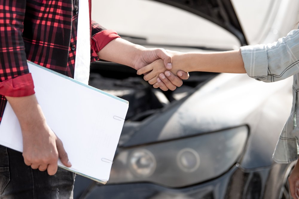 Two individuals shake hands in front of a car with its hood open