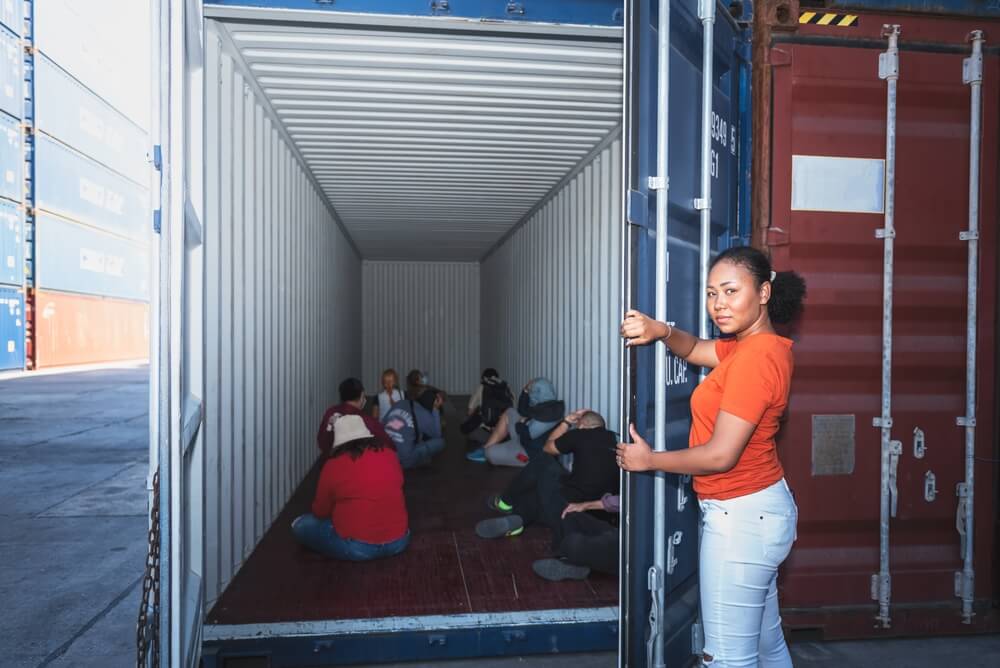 A woman opens a shipping container, revealing several people sitting inside.
