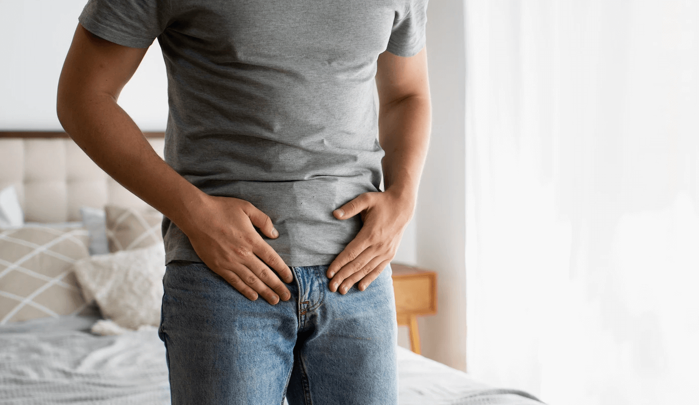 A man in a gray shirt and jeans holds his hands over his groin, appearing to be in discomfort