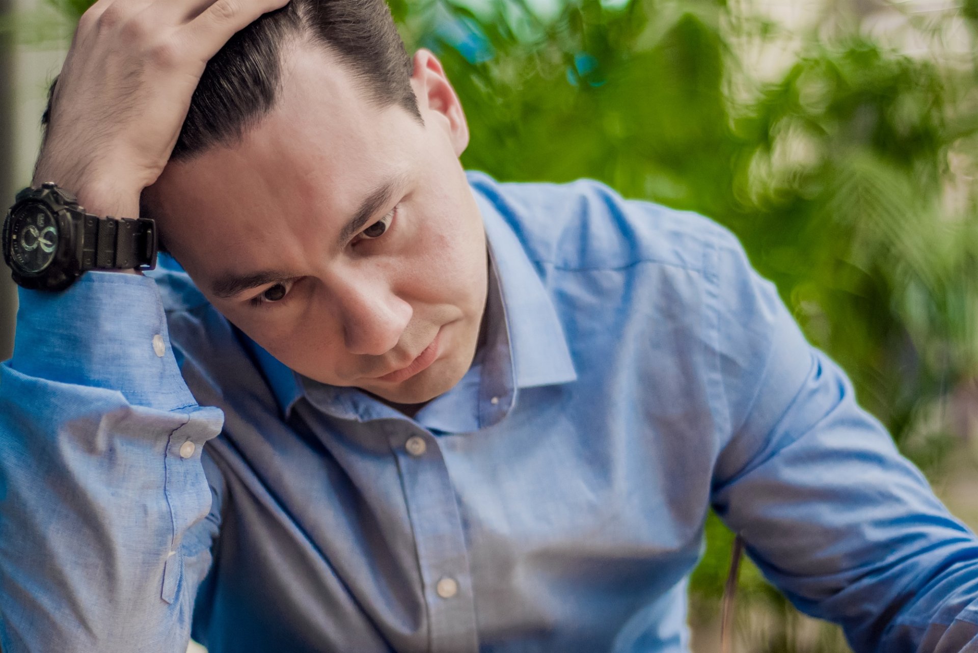 A man in a blue shirt, with his hand on his head, looks down in thought