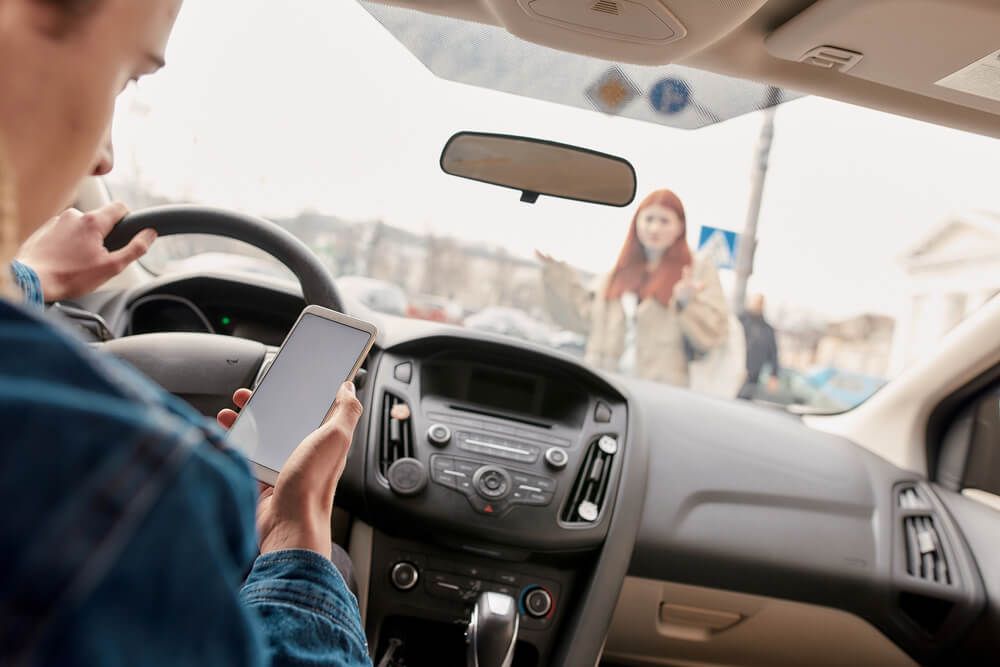 A driver looks at their phone, distracting them from the road and a gesturing person outside the car