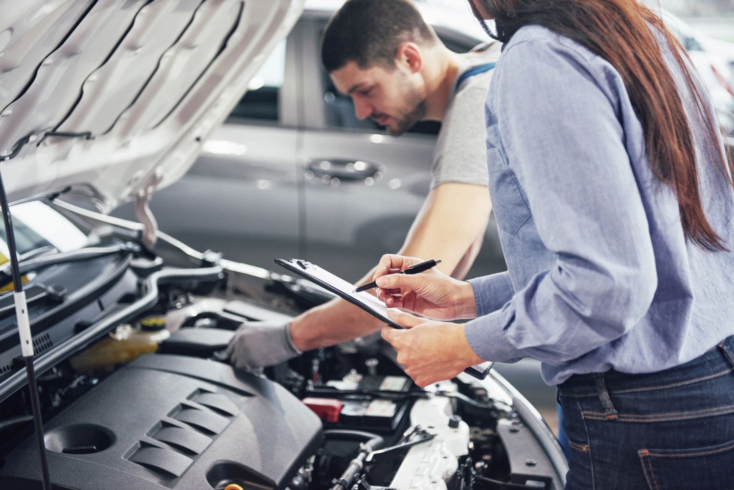 A mechanic inspects a car engine as a woman takes notes on a clipboard, indicating a vehicle safety check.