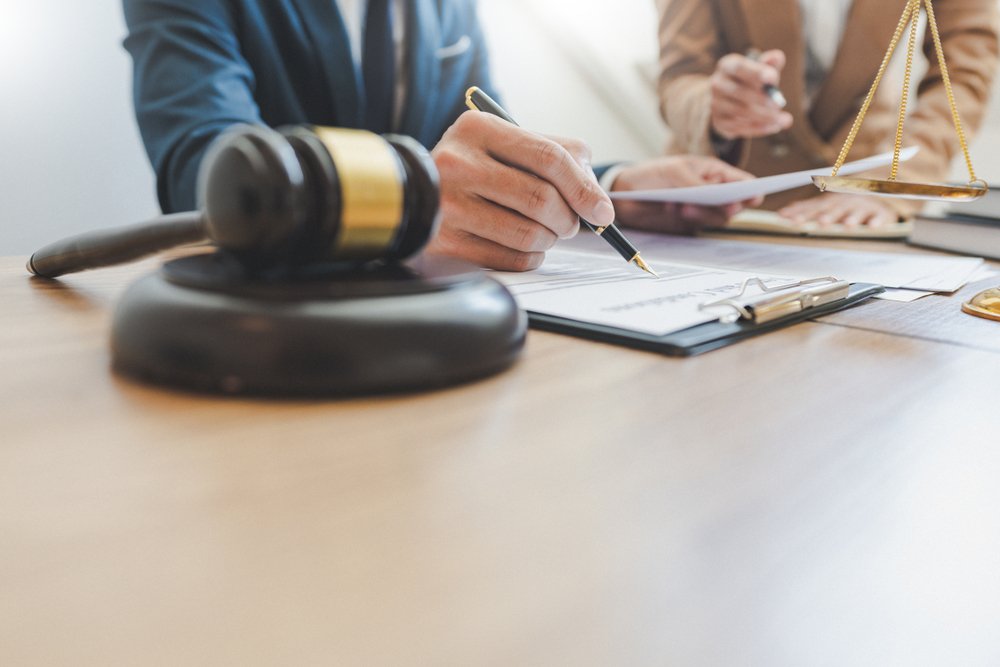 A lawyer signs a document, with a gavel in the foreground and blurred scales of justice on a table