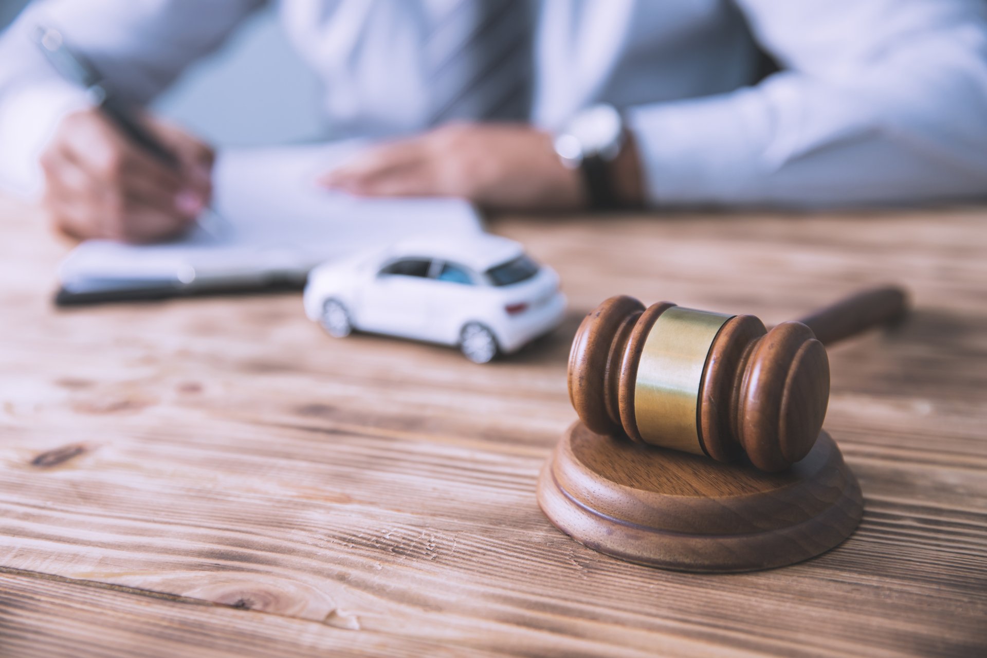 A gavel and miniature car model sit on a wooden desk while a person writes in the background.