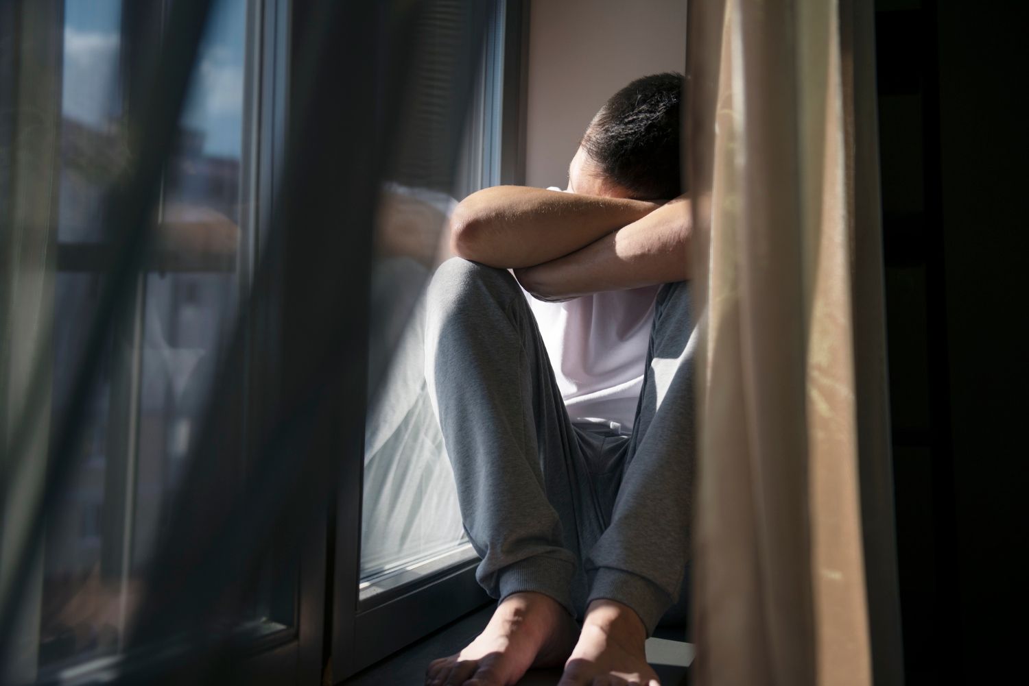 A distressed person, likely a man, sits by a window