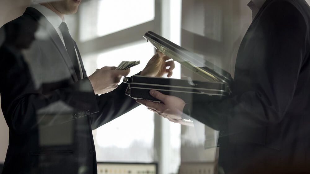 Two men in dark suits exchange money, with one holding stacks of cash and the other an open briefcase.