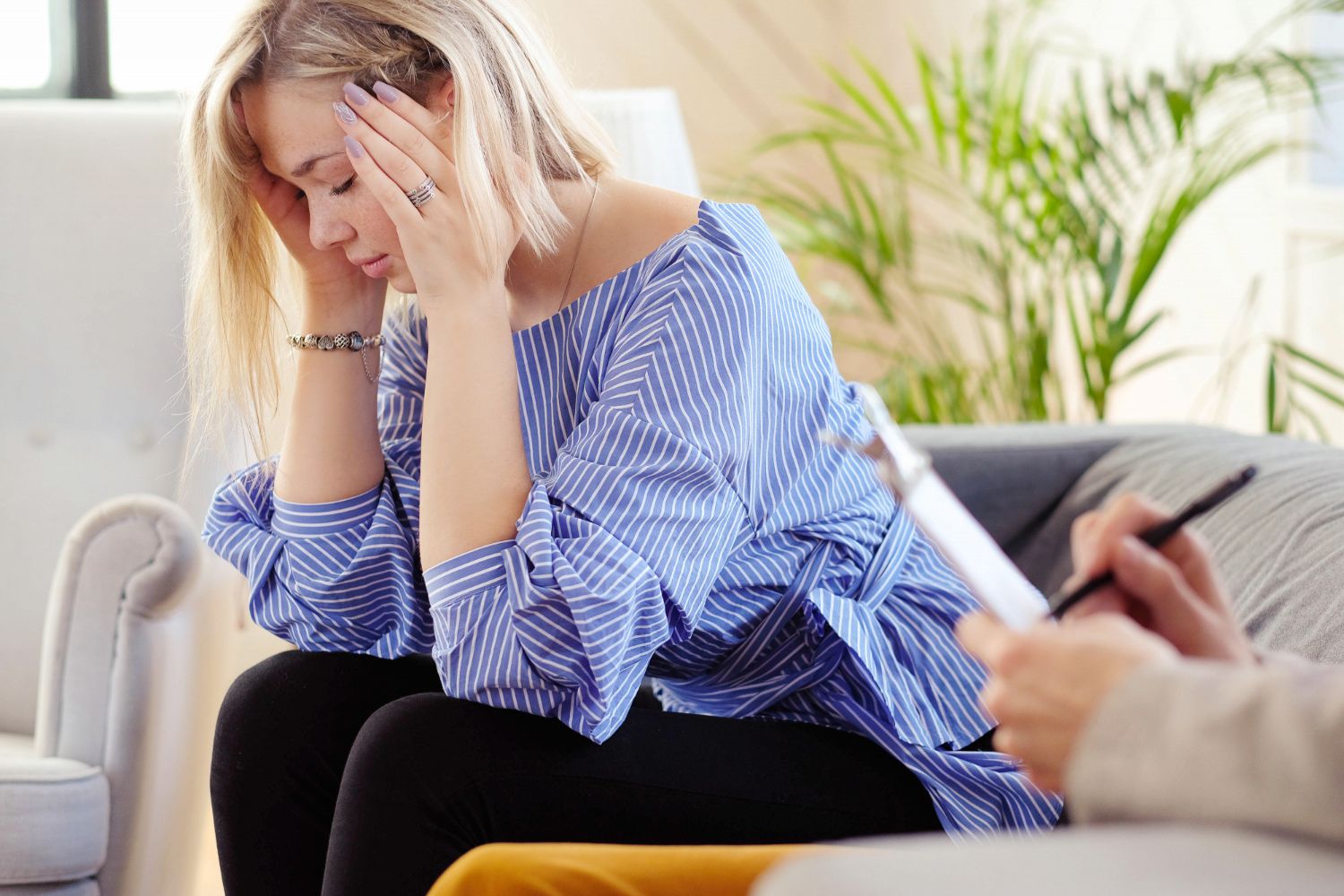 A blonde woman in a blue striped shirt holds her head, eyes closed, appearing distressed
