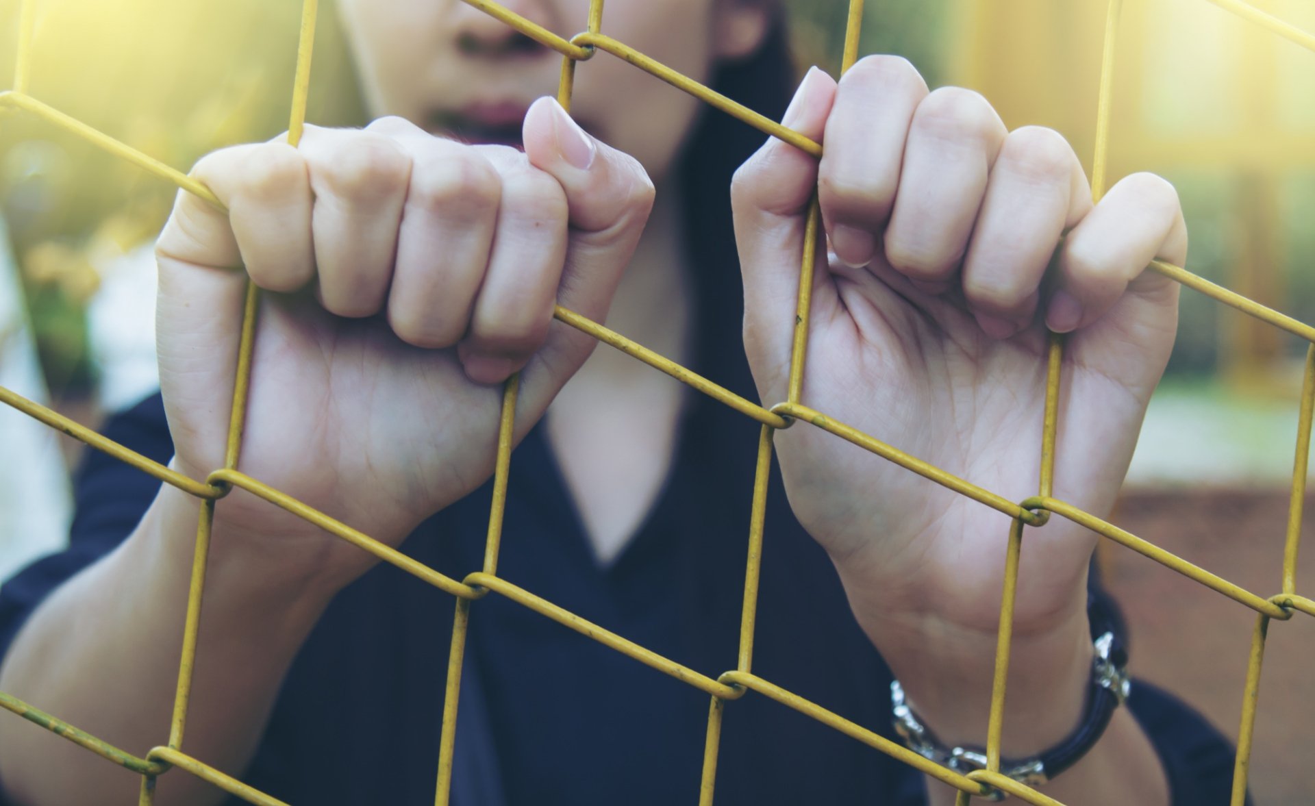 Hands tightly grip a yellow wire fence, symbolizing confinement or imprisonment