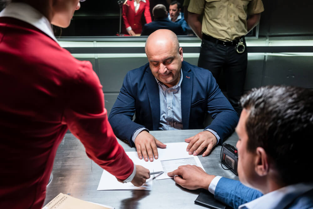 Three people are gathered at a table, with one in red pointing to documents for a man in a dark suit. Another person observes from the side